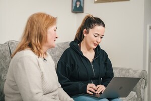 A mother and her adult daughter are sitting on a sofa at home and are looking at a laptop