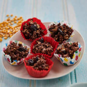 Six rich chocolate and dried fruit clusters nestled in vibrant cupcake liners—red, floral, and polka-dotted—sit on a plate. Packed with oats, chocolate, and chewy dried fruits, their textured surface looks indulgent and satisfying.