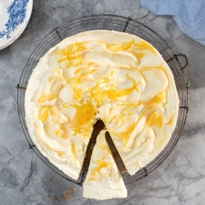 A view of a lemon and ginger cheesecake from above. The cake has a slice cut in it. It is topped with white frosting and yellow drizzle. It is on top of a cake cooling rack.