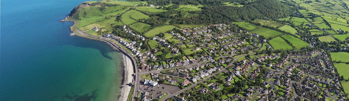 An aerial view of Ballygally in Northern Ireland. It shows several buildings, fields, trees and a coastline.