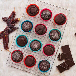 Many muffins are on top of a metal baking cooling rack. The cupcakes are in blue or red cupcake liners. Some of the cupcakes have a sprinkle of white powder on top. Next to the cooling rack are a few red chillies and a broken bar of chocolate. 