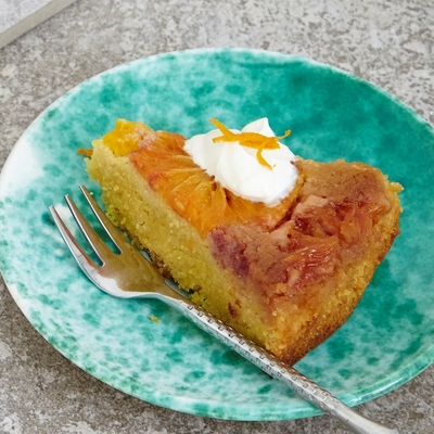 A polenta cake is on top of a square marble plate. A slice has been cut out of the cake. The slice is on a blue patterned plate. It has been topped with cream and an orange garnish. The plate also has a silver fork on it. 