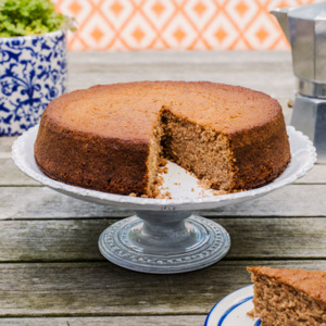 A chestnut cake is on top of a white cake stand. A slice has been cut from the cake. The cake stand is on top of a wooden table. Behind it is a white and blue plant pot and a silver stove top espresso coffee maker. In front of the cake stand is a plate with the cake slice on it. 