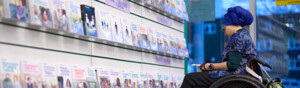 A woman in a wheelchair, wearing a blue headscarf and patterned top looking at a booklet in a healthcare centre. She is in front of a wall displaying cancer booklets.