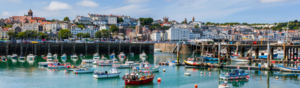The harbour at Saint Peter Port in Guernsey. There are several boats docked in the water to buoys. There are buildings along the harbourside. 