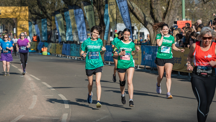 A group of runners are taking part in a race. Some of them are wearing green Macmillan branded sports tops. There are people cheering on either side of the running course.