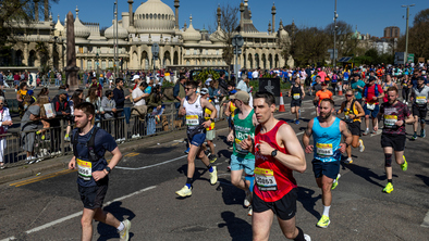 A group of runners are taking part in the Brighton half marathon event. They are wearing running tops, shorts and running shoes. On the side are people cheering for them.