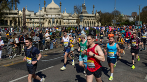 A group of runners are taking part in the Brighton half marathon event. They are wearing running tops, shorts and running shoes. On the side are people cheering for them.