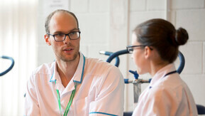 Two Allied Health Professionals wearing white tunics and green Macmillan lanyards sitting and talking to eachother. There are exercise bikes in the background.