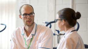 Two Allied Health Professionals wearing white tunics and green Macmillan lanyards sitting and talking to eachother. There are exercise bikes in the background.