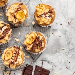 A view of vegan peanut butter and banana cupcakes from above. The cupcakes are placed on a cooling rack. The cupcakes are topped with frosting, chocolate flakes, and baked banana slices. Next to the cooling rack is a broken bar of chocolate. 