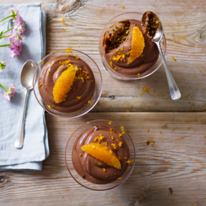A view of vegan chocolate and orange cheesecake pots from above. The pots are placed on a wooden surface. They are decorate with an orange slice and zest. Next to the pots is a blue cotton napkin folded. On top of the napkin is a small spoon and some pink flowers. 