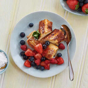 Two cheese blintzes are on a light blue plate. One of the blintzes has been sliced in half. They are topped with strawberries and blueberries, and a white dusting. There is also a silver spoon on the plate. 