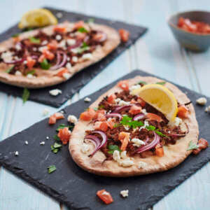 Two lahmacuns are each placed on rectangular slate plates. They are topped with different vegetables and cooked minced meat. 