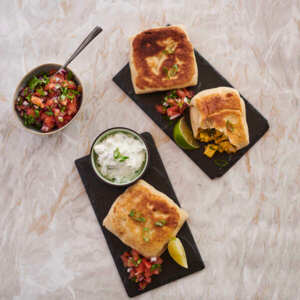 Two dark rectangular slate plates are on top of a light marble table top. On the plates are rotis. On one plate a roti has had a bite taken out of it. Next to the plates is a small bowl with chopped vegetables that has a spoon in it. One plate also has a pot of a white dip in it. 