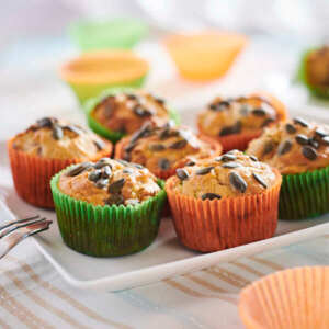 Many muffins are on a white plate. The muffins are in green or orange muffin liners. The muffins are topped with seeds. 