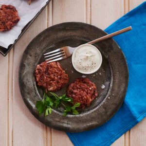 Two beetroot latkes are on a metallic plate. Next to them is a garnish, a pot with a white cream, and a silver fork. The plate is on top of a dark blue cotton napkin and a wooden table. There is also a baking tray with two latkes next to the plate. 