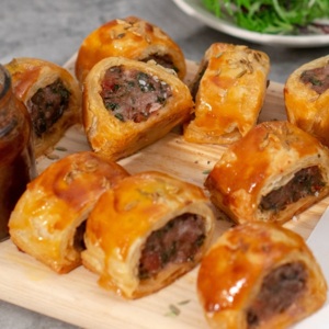Several chorizo sausage rolls are on a wood and plastic board. They are jars of condiments also on the board. Behind them is a plate with different leaf vegetables. 