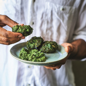 A person in a white shirt is holding a pakroa in one hand and a plate of pakoras in the other. The person is only seen from the shoulders down. The pakroas are green and the plate is a light blue. 