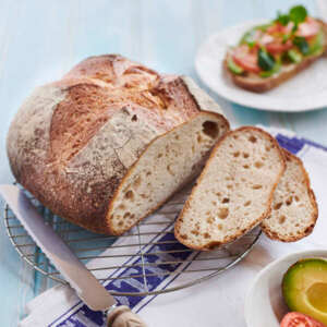A sourdough loaf is on top of a metal cooling rack. The loaf has been cut into slices. Rest on the cooling rack is a metal knife. Underneath the cooling rack is a cotton napkin. Near the loaf is a plate with a slice of bread on it that has been topped with vegetables, and a bowl with a sliced avocado and sliced tomatoes. 