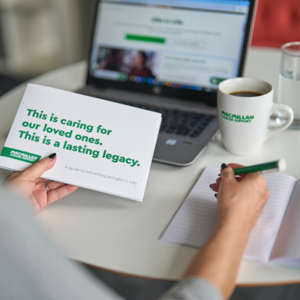 A person is holding a Macmillan Free Wills Service guide in their left hand. In the right hand they are holding a pen over an open notebook that is on a table. In the background is a laptop, a cup of coffee and a glass of water.