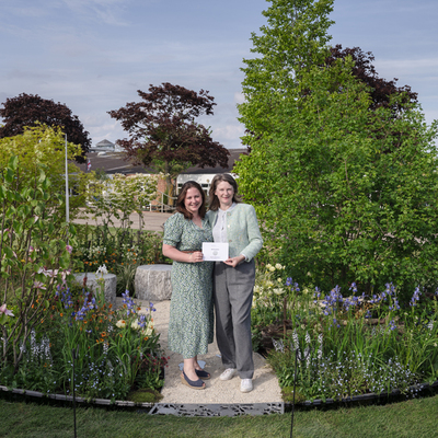 Kerianne Fitzpatrick (left) and TJ Kennedy (right) are standing on a path in the Macmillan Legacy Garden at the at RHS Malvern Spring Festival. They are both holding a piece of paper. The garden is round and had many plants.