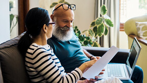 A man and woman sitting on a sofa. They are looking at a document, the man has a laptop on his lap.