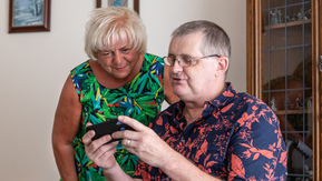 Man in wheelchair is showing the woman standing next to him something on his phone. They're both wearing bright floral tops.