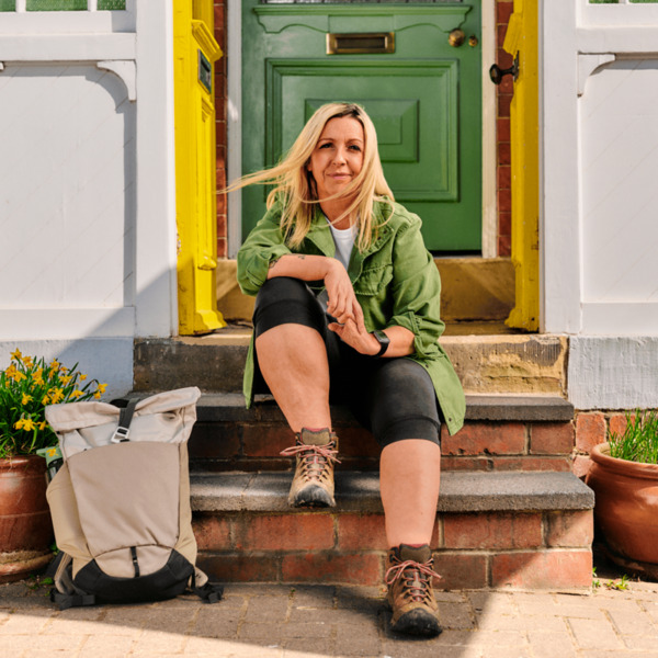 A woman in is sitting on a front door step looking at the camera. She is wearing hiking boots and has a backpack