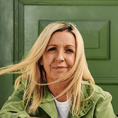 A woman in is sitting on a front door step looking at the camera. She is wearing hiking boots and has a backpack