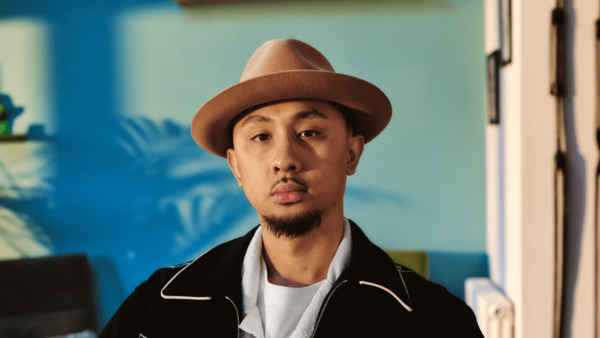 A young man of Asian heritage is sitting at home looking directly to camera. He has a goatee beard and is wearing a hat.