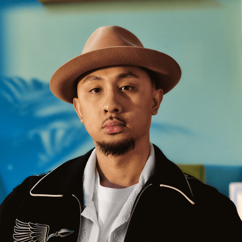 A young man of Asian heritage is sitting at home looking directly to camera. He has a goatee beard and is wearing a hat.