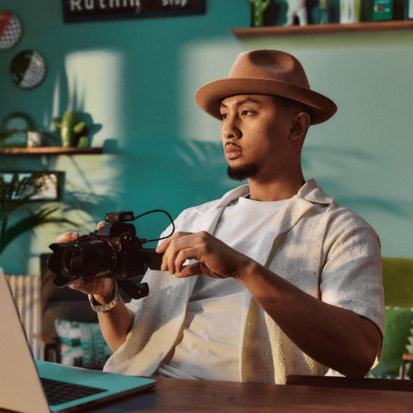 A young man of Asian heritage is sitting at home at a desk in front of a laptop. He's holding a camera. There are plants and ornaments in the background.