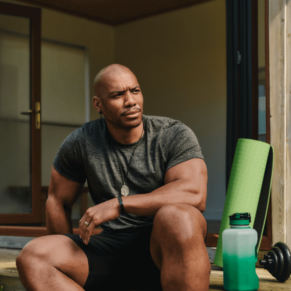 A Black man wearing sports clothes is sitting outside on some decking, He is a body builder and has weights and a water bottle beside him.