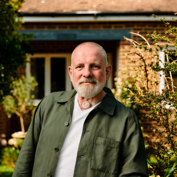 A man is standing outside his home looking to camera. He has a small round valve on his throat.