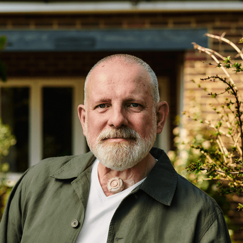 A man is standing outside his home looking to camera. He has a small round valve on his throat.