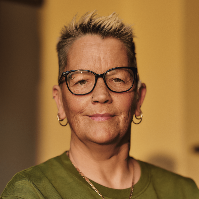 A close-up of a woman looking to camera.  She has short hair and is wearing glasses and has piercings in her ears, nose and eyebrow. Her hearing aid is just visible.