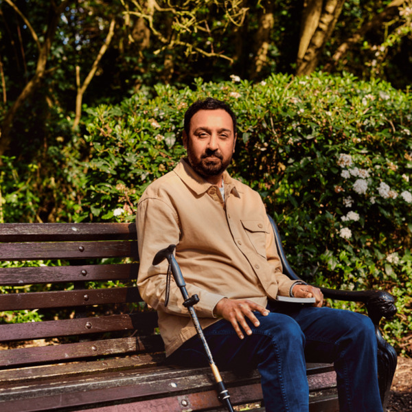 A man is sitting on a park bench. He has a walking stick next to him and a book in his hand.