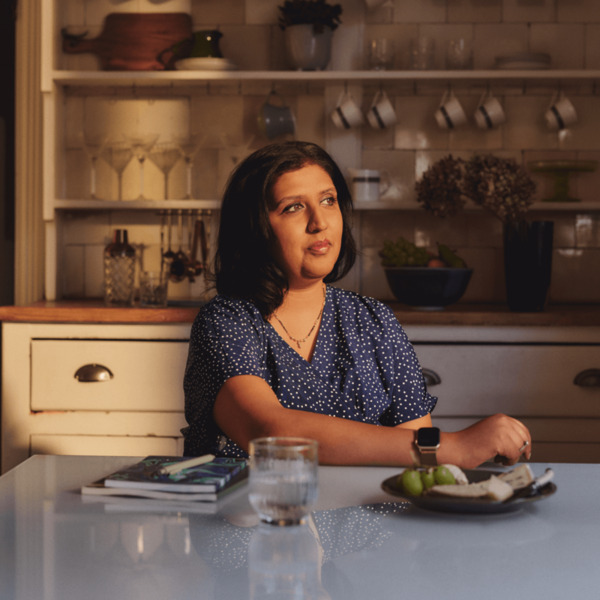 An Asian woman is sitting at a kitchen table looking out of the window. There are notebooks, pens and a plate of food on the table .