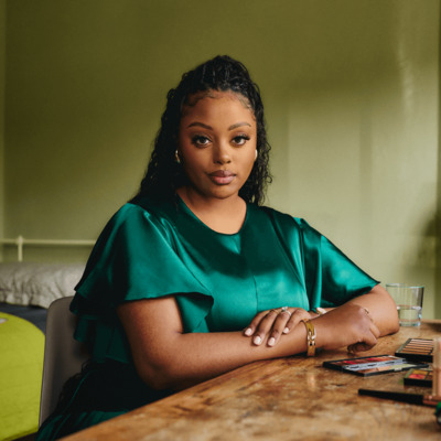 A young Black woman is sitting at home at a table which has make up on. There is a bed in the background with children's toys and books on.