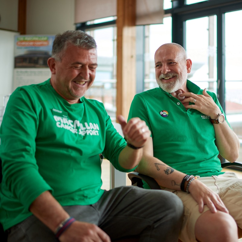 Two volunteers laughing together at the Macmillan Horizon Centre wearing Macmillan shirts. One man is holding a valve on his throat. 