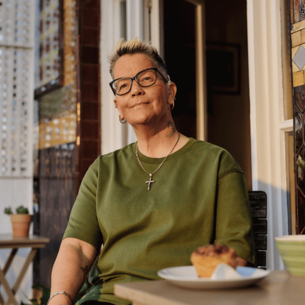 A woman is sitting at a patio table with a coffee and cake. She is wearing glasses and a hearing aid.
