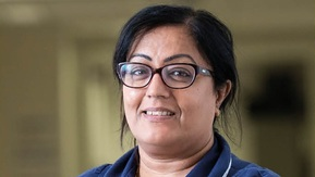 A Macmillan healthcare professional smiling at the camera wearing glasses and a dark blue uniform, holding a stack of booklets.