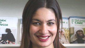 Azmina, a Macmillan Recovery Package Project Manager, smiling in front of a shelf of Macmillan booklets.