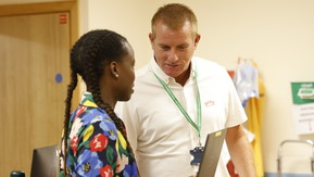 Two healthcare professionals in conversation. The man is wearing a white shirt and the woman is holding a laptop, she is wearing a bright floral dress.