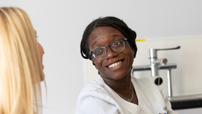 Two healthcare professionals in conversation. The woman in the centre is smiling and looking toward the other, wearing glasses. There are papers, a phone, and a pen on the table. 