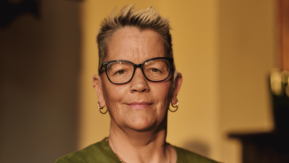 A close-up of a woman looking to camera.  She has short hair and is wearing glasses and has piercings in her ears, nose and eyebrow. Her hearing aid is just visible.