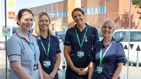 A group of 4 nurses standing outside a hospital. Three are wearing dark blue uniforms and one is wearing a light blue uniform.