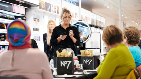 A Boots Beauty Advisor is standing and speaking to a group of people sitting in chairs. They are inside a Boots store. On the table in front of the group are bags with No7 branding on them.