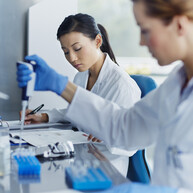 Two female scientists working in a laboratory. One is using a pipette to handle a sample, while the other writes notes on a document. 